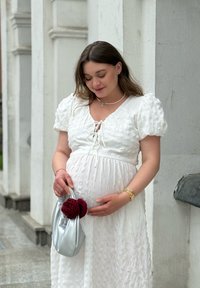 Gravid kvinna i vit klänning som varsamt håller om sin mage, bär en silverväska med en röd blomma, stående bredvid vita byggnadssäulen.