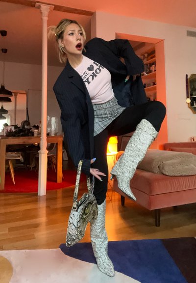Mujer con blazer, minifalda y botas con estampado levantando una pierna, sosteniendo un bolso a juego en una sala de estar con luz cálida.