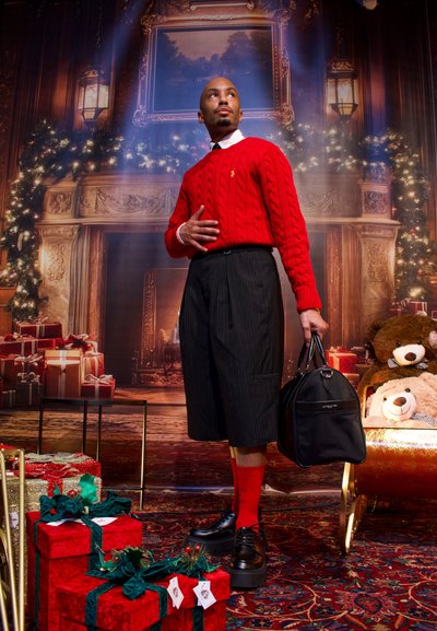 Jersey rojo de punto con cable, pantalones cortos negros a rayas, zapatos negros y calcetines rojos. Sosteniendo un bolso negro, con regalos navideños y osos de peluche de fondo.