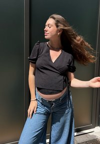 Černý crop top s krátkými rukávy, textura látky, v kombinaci se světle modrými džínami s volnými nohavicemi. Viditelný je těhotenský pupík a doplňky.