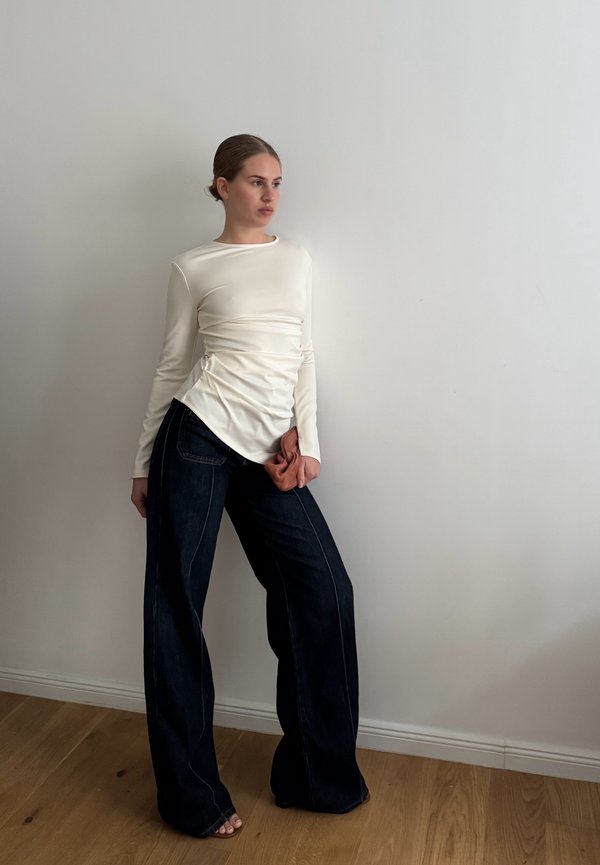 Haut crème à manches longues avec un détail froncé à la taille, associé à un jean large taille haute en denim noir. Sol en bois et mur blanc en arrière-plan.