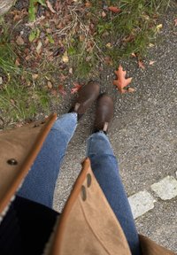 En person som har på seg blå jeans, brune lær støvler og en tan farge frakk, står på en grusvei med falne høstblader og gress i nærheten.