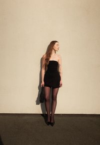 Haut noir sans bretelles avec un ourlet volants, associé à un short noir. La silhouette porte des collants noirs transparents et des escarpins noirs à bout pointu, tenant un sac noir.