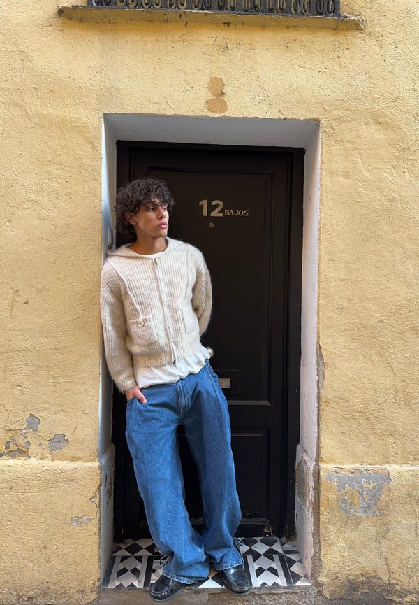Young person with curly hair wearing a cream jumper and loose blue jeans stands leaning against a black door numbered 12 Bajos.