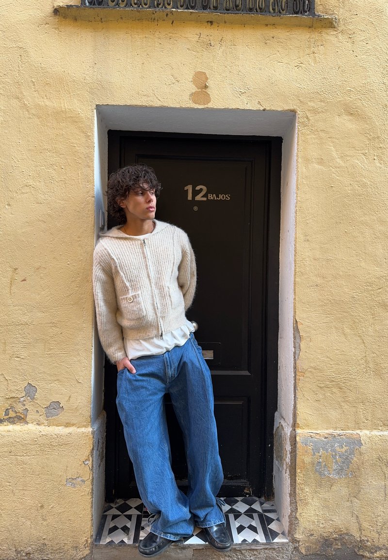 Jongere met krullend haar, gekleed in een crèmekleurige trui en ruime blauwe spijkerbroek, staat tegen een zwarte deur met nummer 12 Bajos geleund.