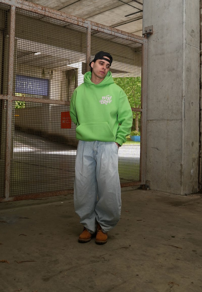Jonge man in een groene hoodie en wijde jeans staat binnen bij een metalen poort, draagt een zwarte pet achterstevoren en bruine laarzen.