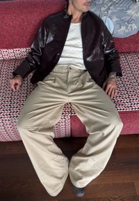 Brun glanset jakke, hvit strikket topp, beige vide bukser, sitter på en mønstret sofa med rødt og hvitt geometrisk design.