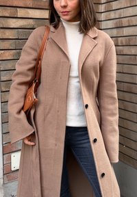 Manteau en laine beige clair avec col, boutonné à l'avant, accompagné d'un col roulé blanc côtelé et d'un jean bleu. Conçu avec des manches larges.