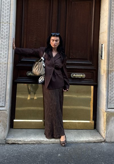 Blazer marrón sobre una falda maxi marrón con estampado, combinado con un bolso gris. Puerta de madera texturizada con herrajes de latón al fondo.