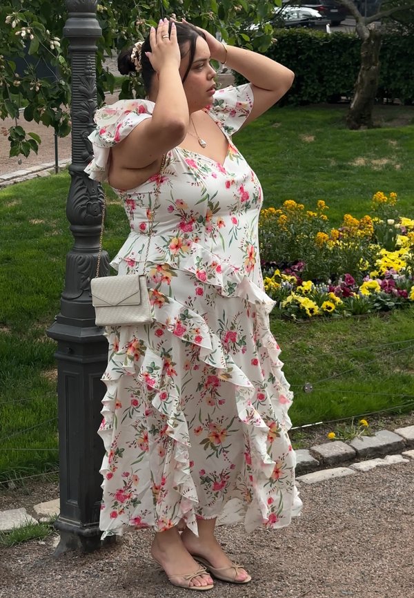 Frau in einem geblümten Rüschkleid steht auf einem Gartenweg, hält ihren Kopf mit beiden Händen nahe einem Laternenpfahl und blühenden gelben Blumen.