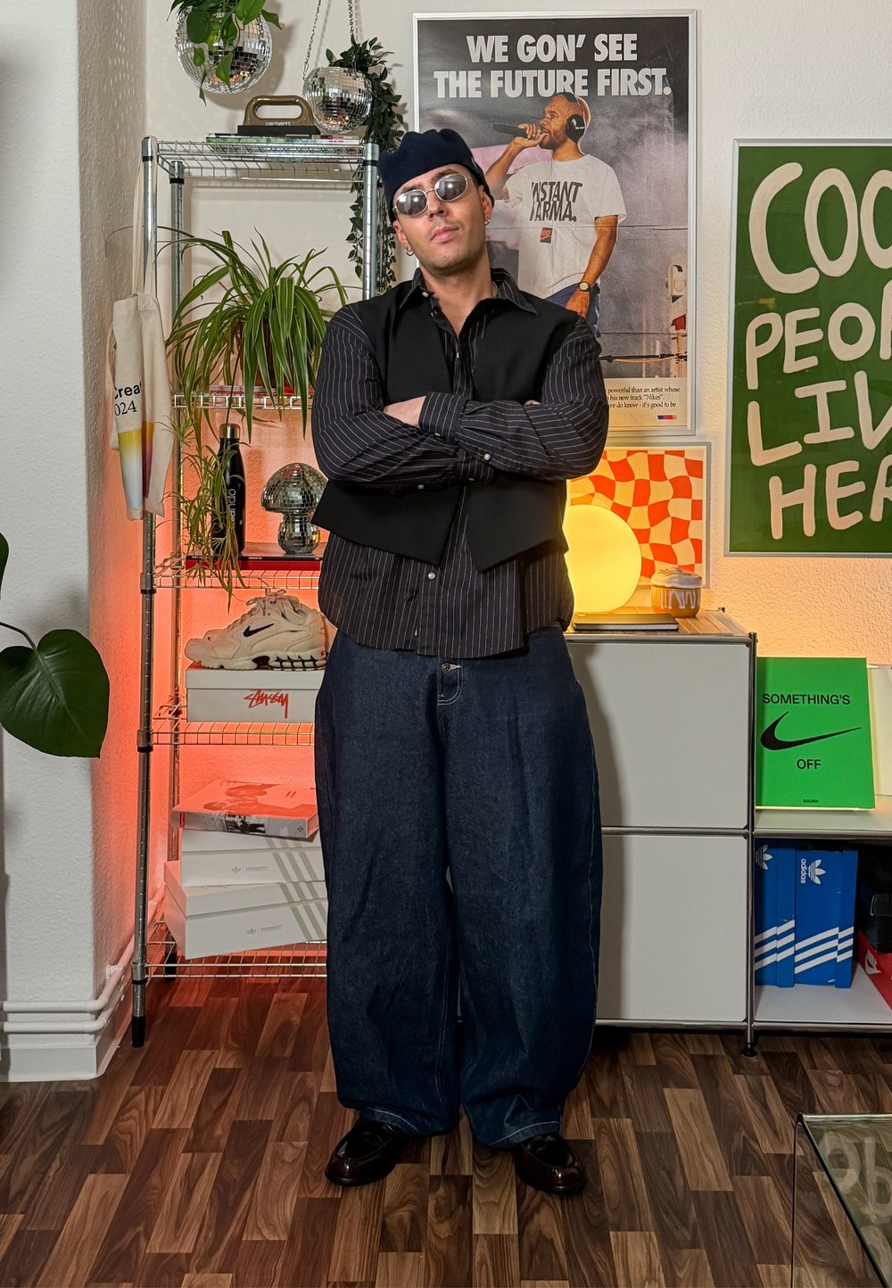 Homem de óculos de sol, boina, camisa às riscas e colete está de braços cruzados numa sala decorada com cartazes e plantas.