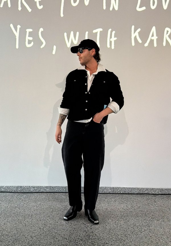 Homme portant une casquette noire, des lunettes de soleil, une veste noire et un pantalon foncé, debout à l’intérieur contre un mur blanc avec un texte partiel visible au-dessus.
