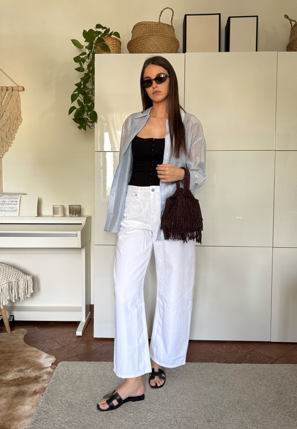 Femeie care poartă top negru, pantaloni albi largi, cămașă deschisă cu dungi, ochelari de soare, ținând o geantă maro cu franjuri într-o cameră luminoasă cu plante și pian.