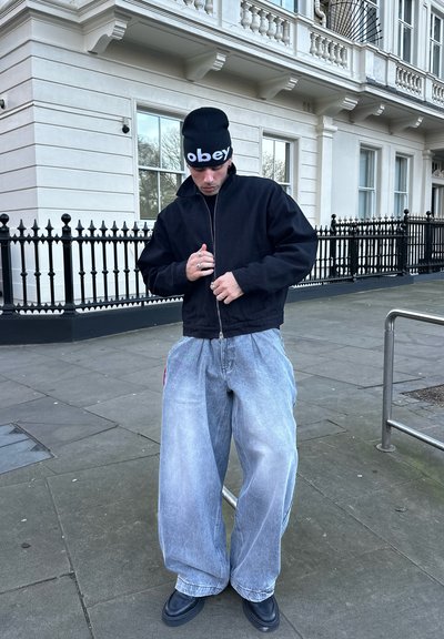 Joven con gorro negro, chaqueta negra, vaqueros azules claros de gran tamaño y zapatos negros, de pie en una calle de la ciudad ajustando la cremallera de su chaqueta.
