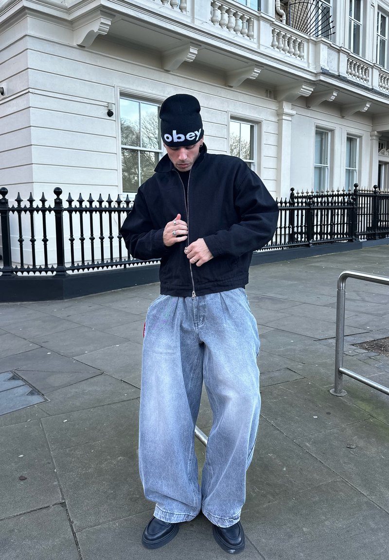 Jeune homme portant un bonnet noir, une veste noire, un jean bleu clair oversize et des chaussures noires, debout dans une rue de la ville en train d'ajuster la fermeture éclair de sa veste.