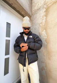 Μαύρο φανελένιο μπουφάν με γυαλιστερή επιφάνεια, με υψηλό γιακά, τσέπες με φερμουάρ και λογότυπο στην αριστερή πλευρά του στήθους. Συνδυασμένο με κρεμ παντελόνια.