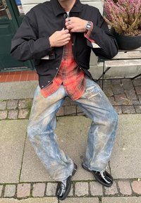 Personne assise à l'extérieur sur un banc portant une veste noire, une chemise à carreaux rouge, un jean large déchiré, des bagues, une montre digitale et des chaussures noires.