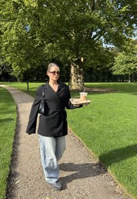 Svart omslag blazer, lyseblå vidbenede jeans og solbriller. Holder en kaffe i en papplatet mens han/hun går på en grussti gjennom grønne områder.