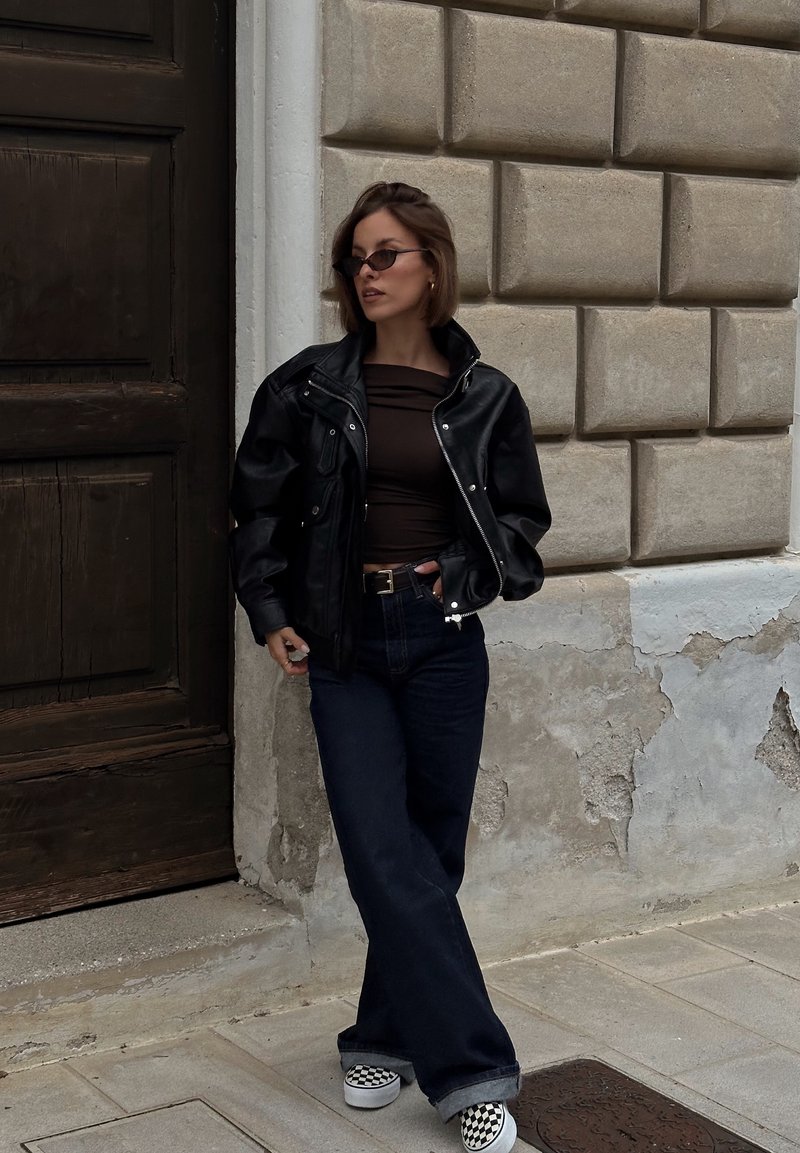Black leather jacket, brown fitted top, dark blue wide-leg jeans, and black-and-white checked slip-on shoes. Standing against a textured wall.
