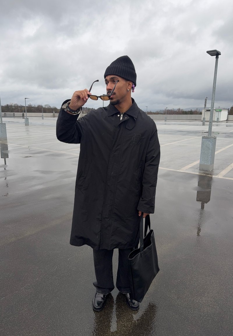 Uomo con cappotto nero e berretto regolando gli occhiali da sole, con una borsa tote nera, in piedi su un parcheggio sul tetto bagnato sotto un cielo nuvoloso.