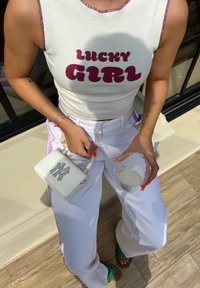 Λευκό ριμπ κορμάκι με το γράμμα "LUCKY GIRL" σε μωβ, λευκά παντελόνια ψηλής μέσης, μικρή λευκή τσάντα με ριχτίνες, διάφανο ποτήρι, πολύχρωμες σαγιονάρες.