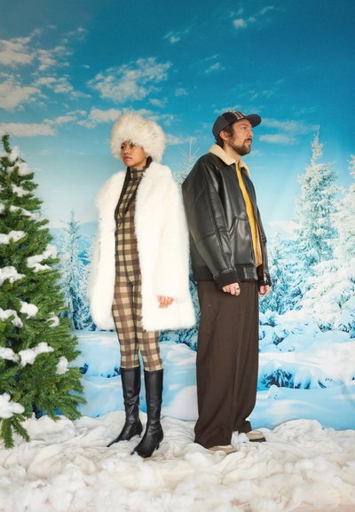 Mujer con abrigo y sombrero de piel blanca, traje de cuadros y botas, de pie junto a un hombre con chaqueta de cuero y pantalones anchos marrones, con un fondo de árboles nevados.