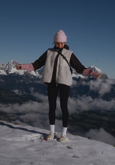 Chaleco forrado de forro polar en gris, superpuesto sobre una blusa negra con mangas rosas, combinado con leggings negros y zapatillas coloridas sobre la nieve.