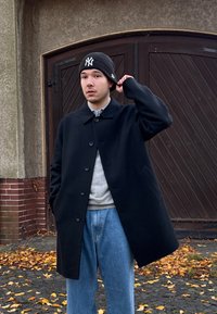 Juodas vilnonis paltas su apykakle, šešių mygtukų u�žsegimas ir šoninės kišenės, dėvimas ant pilko megztinio ir šviesiai mėlynų džinsų. Juoda medvilninė kepurė.