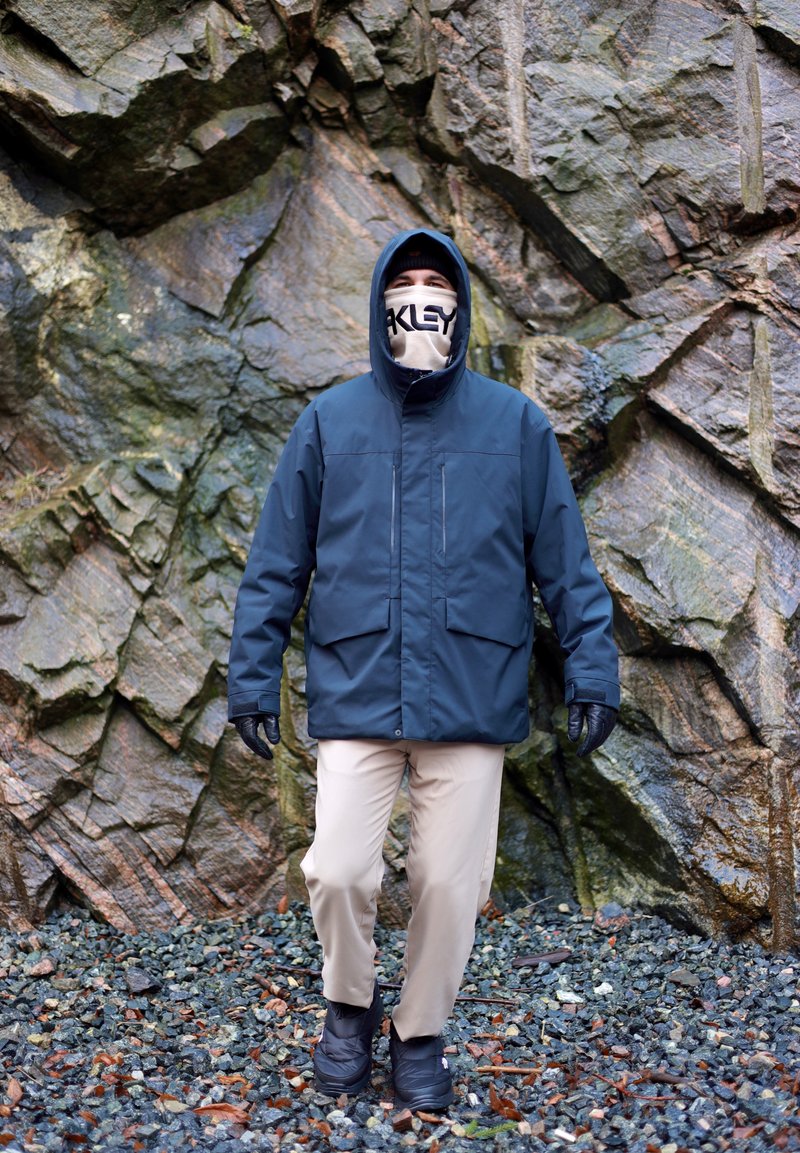 Marineblaue wasserdichte Jacke mit Kapuze, zwei Vordertaschen, schwarzen Handschuhen, beigen Hosen und schwarzen Stiefeln vor felsigem Gelände.