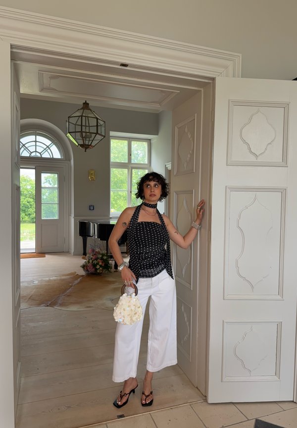 Femeie cu maiou negru cu buline și pantaloni albi cu picioare largi stă în ușă, ținând o geantă albă florală într-o cameră luminoasă și elegantă.