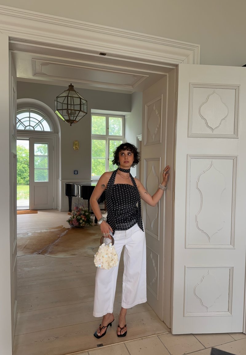 Femme en haut dos nu noir à pois et pantalon blanc à jambes larges se tient dans l'encadrement de la porte, tenant un sac à main blanc à fleurs dans une pièce lumineuse et élégante.