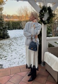 Femme en pull bleu clair en tissu duveteux, jupe blanche à pois avec dentelle noire, bottes noires et sac matelassé bleu marine, debout sur une terrasse enneigée près d'une couronne.