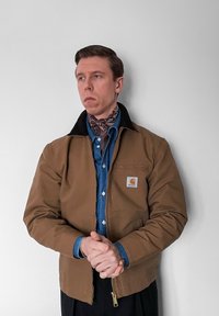 Veste en toile marron avec un col noir, arborant un logo sur la poche poitrine, superposée à une chemise en denim bleu, portée avec une écharpe marron à motif.