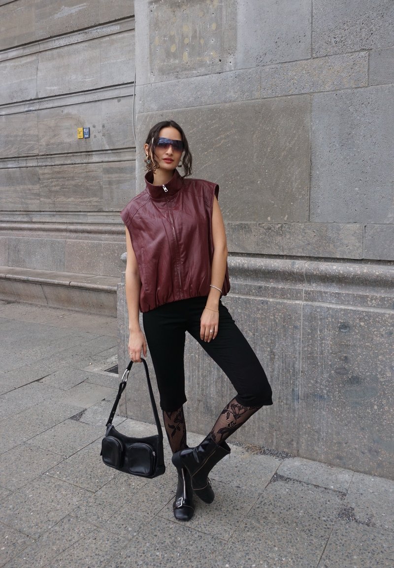 Vrouw draagt een mouwloze bordeaux leren top, zwarte cropped broek, patroon kousen, zwarte laarzen, zonnebril en houdt een zwarte handtas tegen een stenen muur.