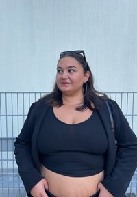 Μαύρο ριμπ crop τοπ κάτω από ένα μαύρο σακάκι; το μοντέλο έχει μακριά μαλλιά, σκουλαρίκια κρίκους και εμφανή υφή στο δέρμα. Φόντο σε ανοιχτό γκρι.