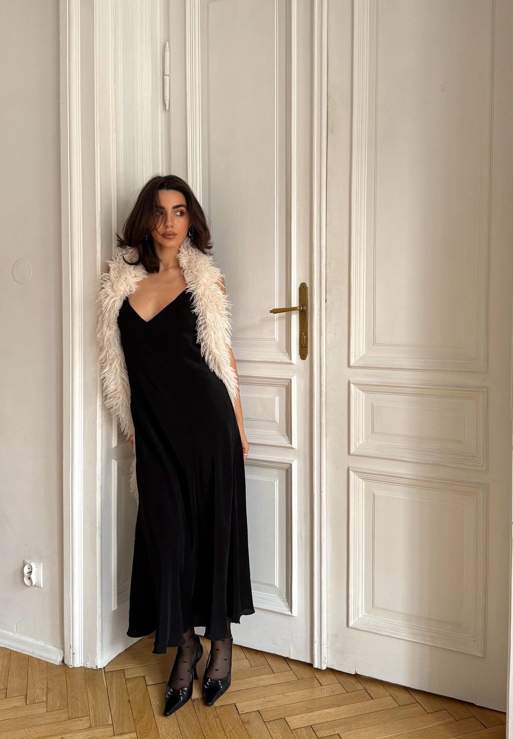 Robe noire sans manches au design fluide, associée à un châle en fausse fourrure blanc. La tenue est complétée par des bas à pois et des escarpins noirs.