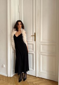 Robe noire sans manches au design fluide, associée à un châle en fausse fourrure blanc. La tenue est complétée par des bas à pois et des escarpins noirs.
