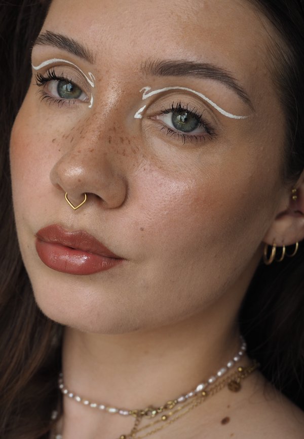 Gros plan sur une femme avec de légères taches de rousseur, un anneau de septum en or, un eye-liner blanc au-dessus des yeux, un maquillage naturel et des colliers superposés en or et perles.