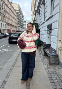 Femme aux longs cheveux portant un pull crème et rouge, un jean à jambes larges et des chaussures marron, tenant un sac tissé sur un trottoir dans une ville européenne.