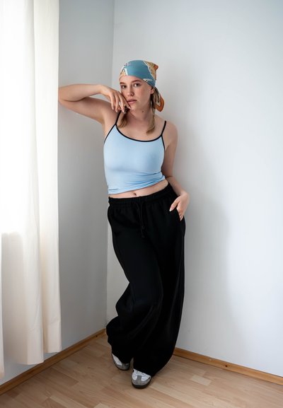 Top sin mangas azul claro con ribetes negros, combinado con pantalones de chándal negros holgados. Una bufanda de seda estampada cae sobre su cabello. Pared neutra y suelo de madera.