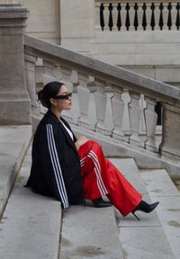 Blazer noir oversize avec des rayures blanches, pantalon rouge à jambes larges avec des rayures blanches, talons hauts noirs, et lunettes de soleil, assis sur des marches en pierre.