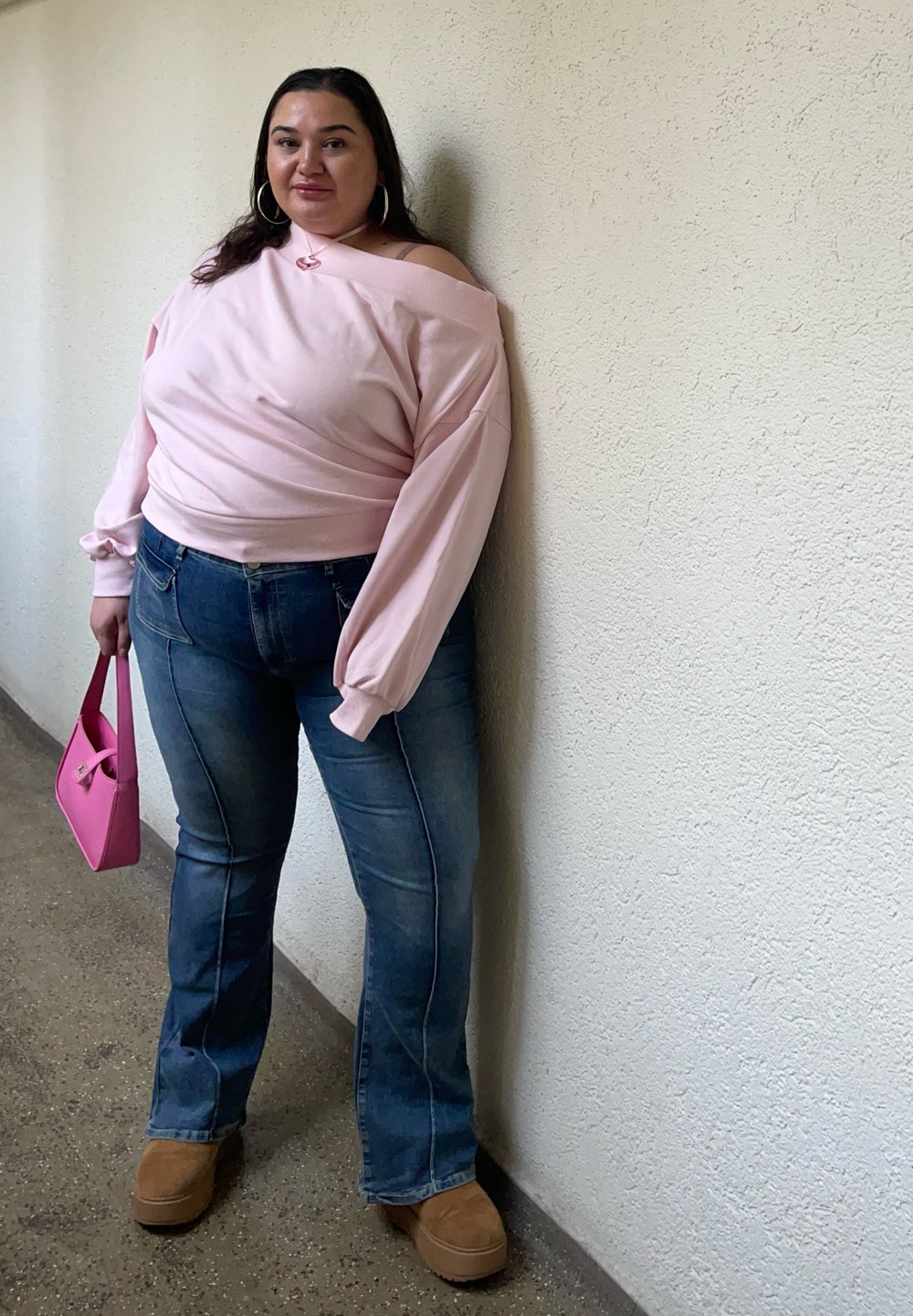 Sweatshirt rosa de ombros descobertos, jeans azuis de perna larga, botas de tom castanho e uma pequena bolsa rosa. Texturas suaves com costuras e detalhes de pontos visíveis.