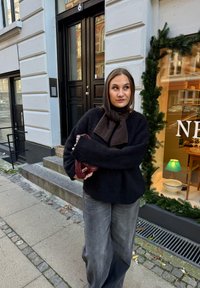 Čierny nadýchaný sveter so širokými rukávmi, sivé široké nohavice a tmavohnedý šál. Drží zapletenú bordovú tašku. Mestské pozadie.