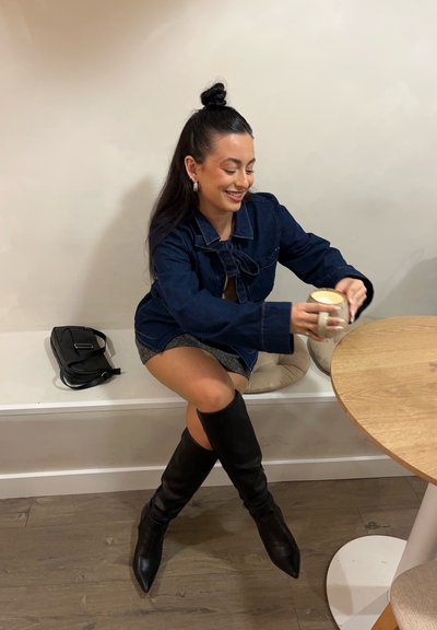 Chaqueta de mezclilla con detalle de pajarita, falda gris, botas negras hasta la rodilla, sosteniendo una taza blanca en una mesa de madera, bolsa negra a su lado.