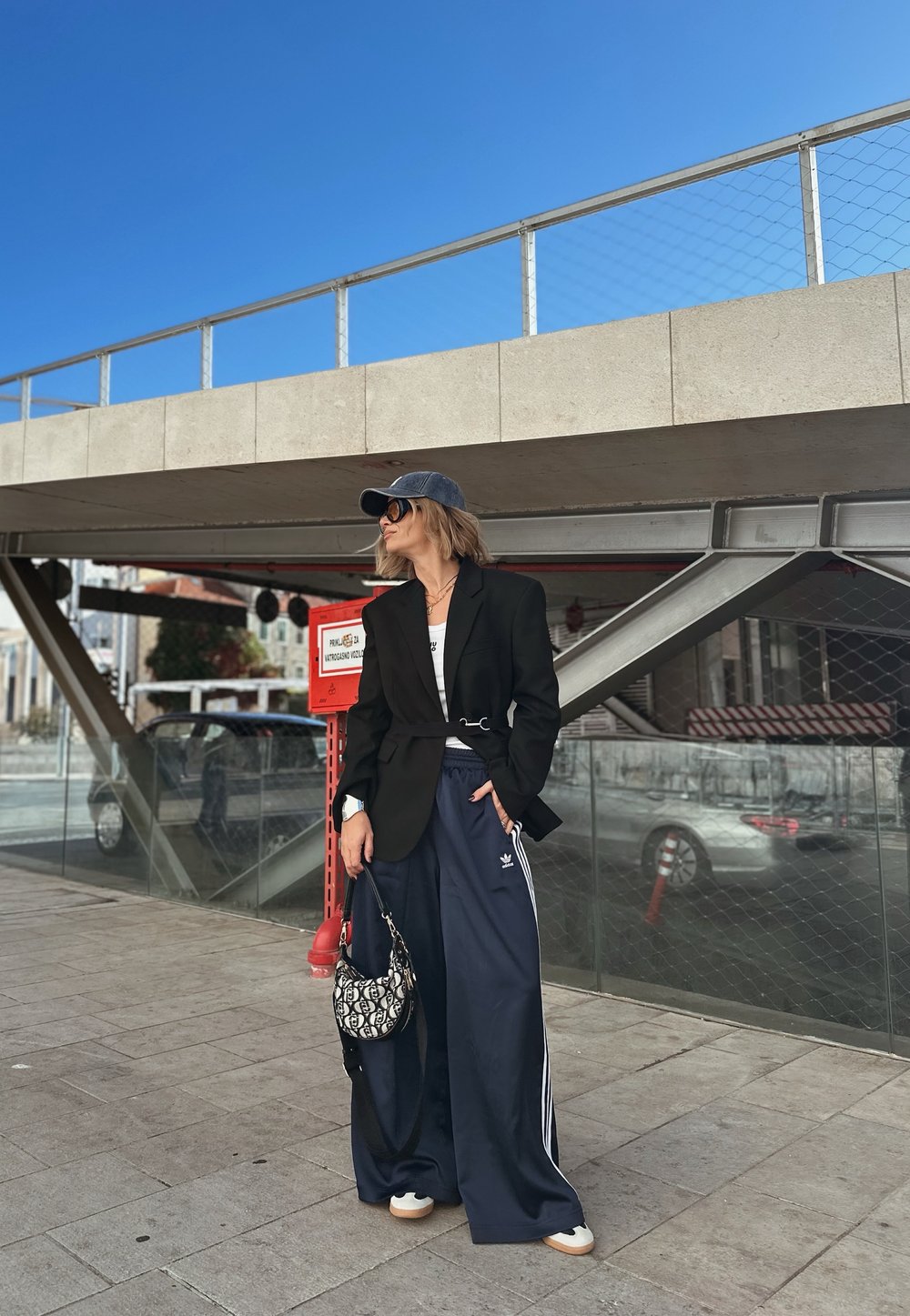 Nero giacca oversize, t-shirt bianca, pantaloni larghi blu navy con strisce bianche, cappello grigio, occhiali da sole neri e borsa a fantasia.