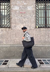 Homme portant une casquette noire et des lunettes de soleil, une chemise à motifs, un pantalon noir large, et portant un grand sac noir marchant devant un mur en pierre avec des fenêtres grillagées.