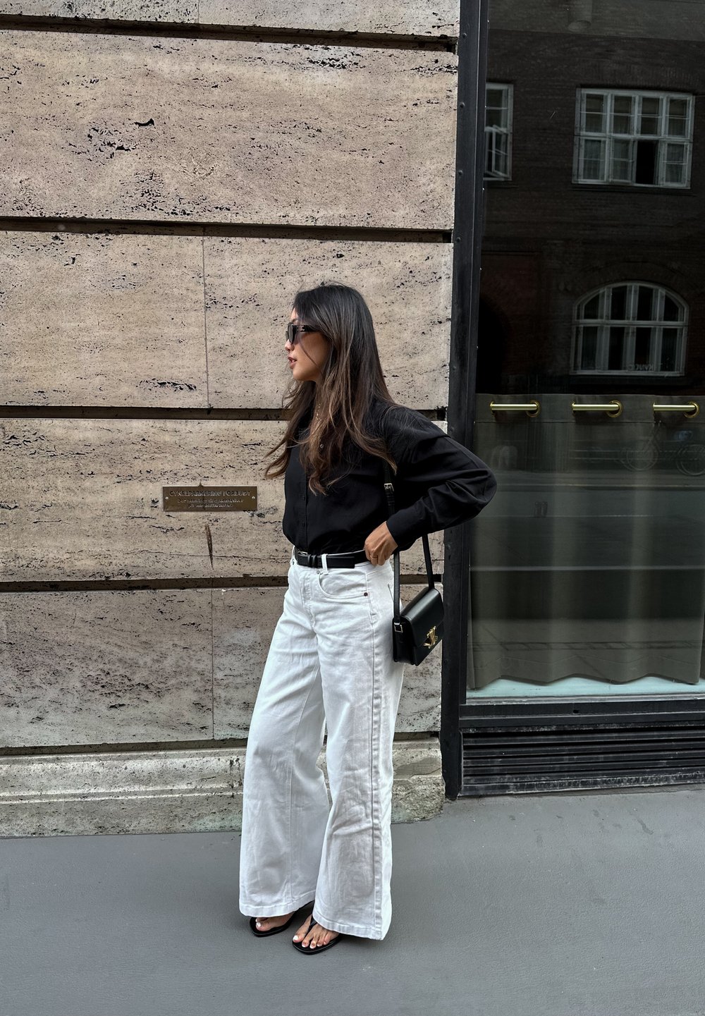 Sort skjorte med knapper, brede hvide jeans, sort bælte, sort crossbody taske og sorte sandaler. Langt hår, står op ad en tekstureret væg.