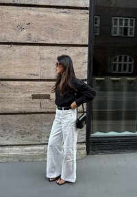 Sort skjorte med knapper, brede hvide jeans, sort bælte, sort crossbody taske og sorte sandaler. Langt hår, står op ad en tekstureret væg.