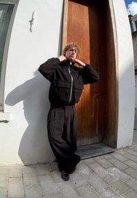 Μαύρο φλις μπουφάν με ψηλό γιακά και μπροστινές τσέπες, σε συνδυασμό με μαύρο, φαρδύ παντελόνι. Η σκηνή περιλαμβάνει μια ξύλινη πόρτα.