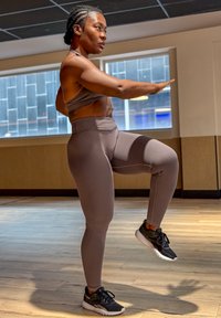 Mujer con ropa deportiva equilibrándose sobre una pierna con los brazos extendidos en el interior, sobre un suelo de madera cerca de grandes ventanas.